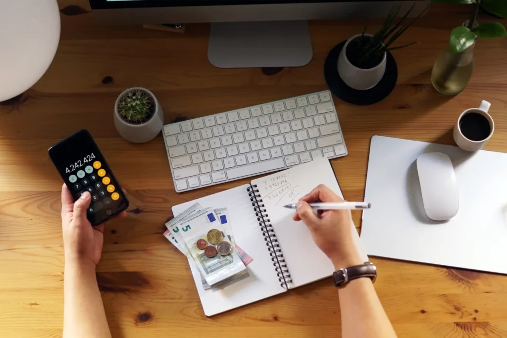 Imagem de uma mesa de trabalho com computador, caderno, calculadora, dinheiro em euros, caneta, planta e xícara de café, símbolo de finanças pessoais e controle financeiro.